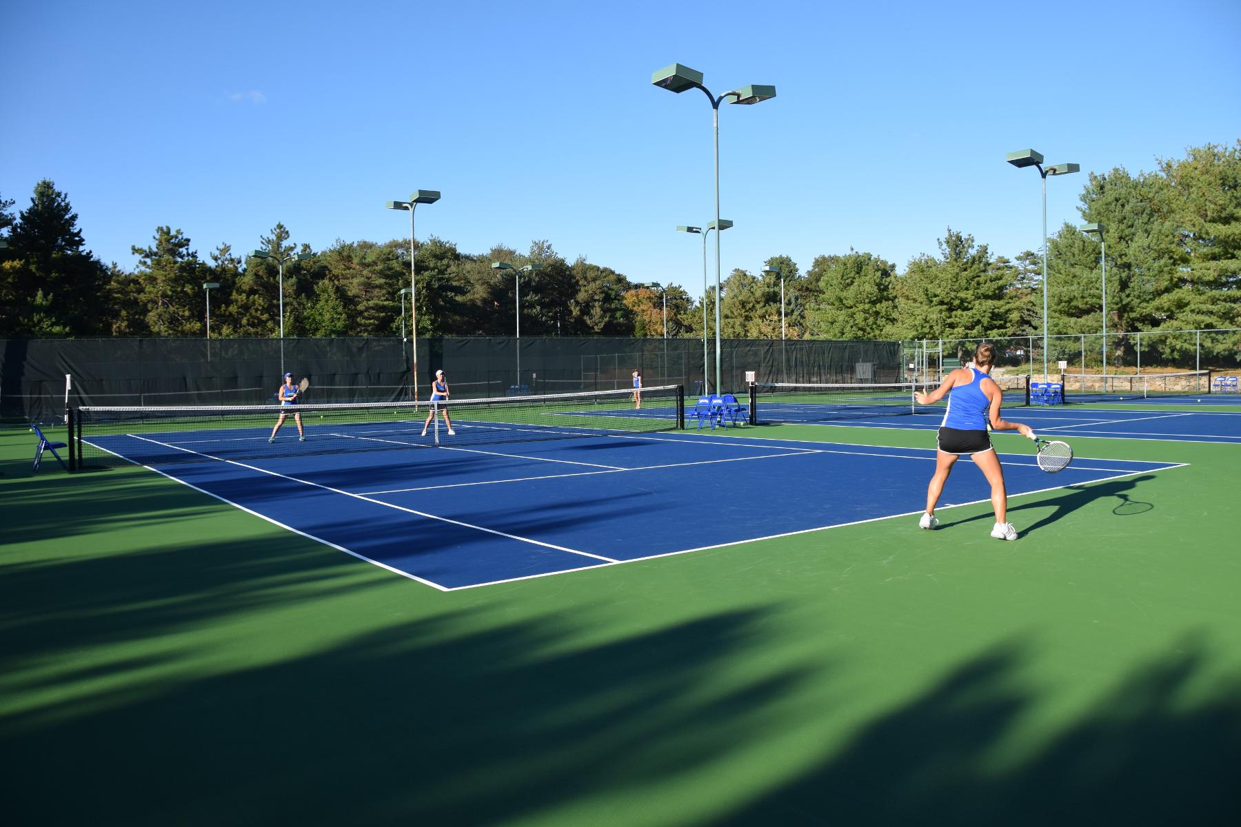 Panoramic view of outdoor tennis courts with athlete in the foreground hitting from the baseline 